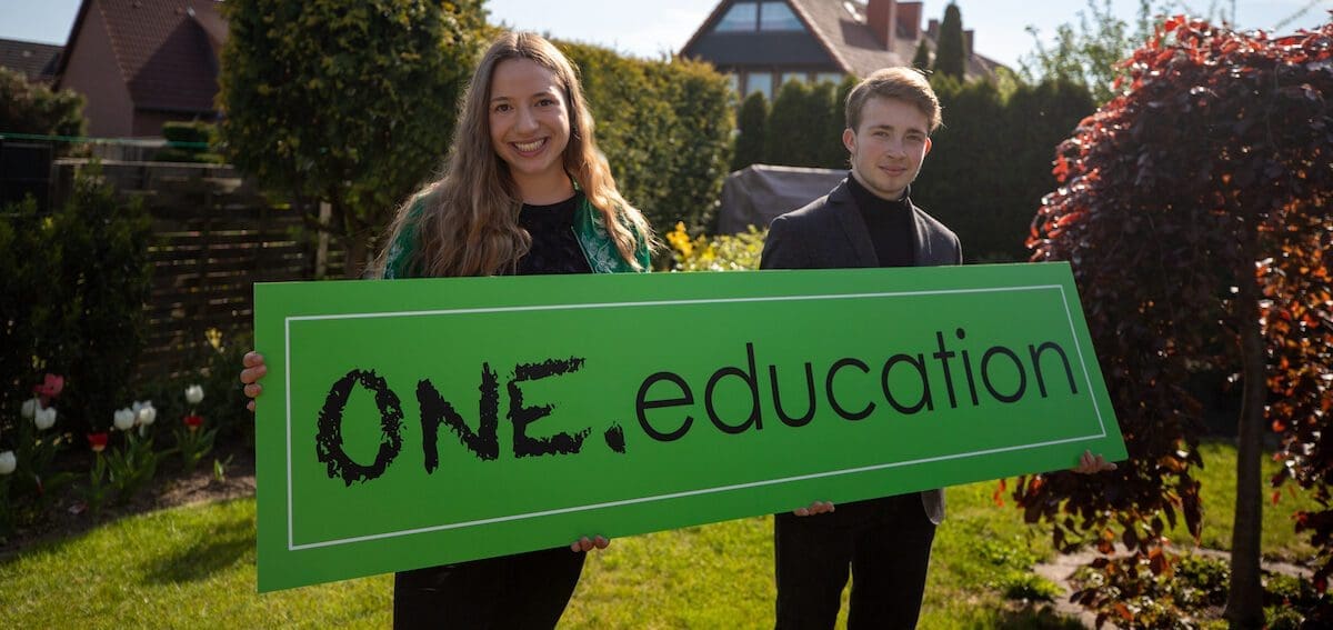 Die Gründerin Laura und der Gründer Joscha stehen im Garten und halten ein großes Schild mit dem Namen ihres Startups One.Education hoch.
