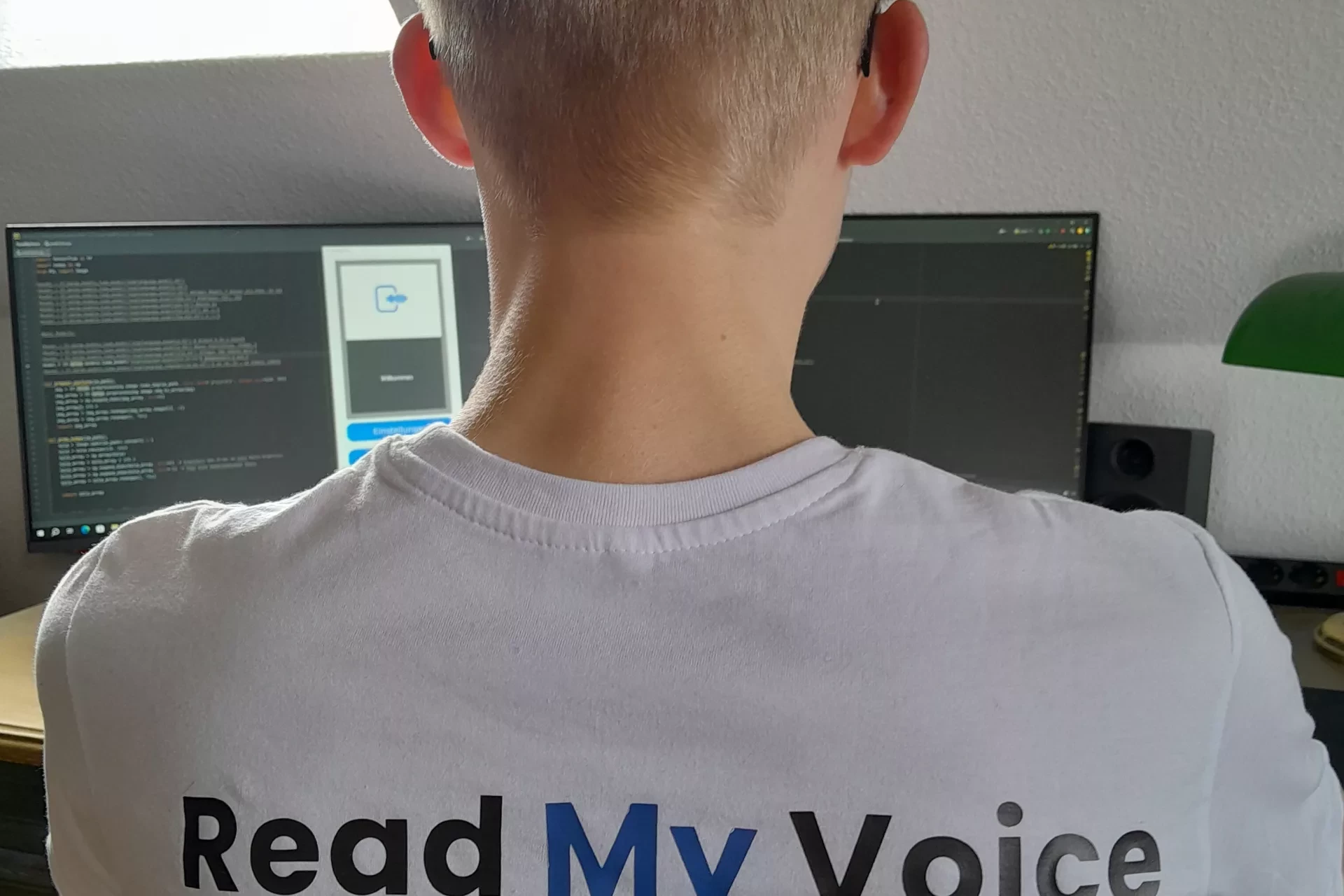 Der Gründer Mika Danner vor seinem Computer von hinten fotografiert. Auf dem Rücken seines T-Shirts ist das Logo seines Startups Read my Voice zu sehen.